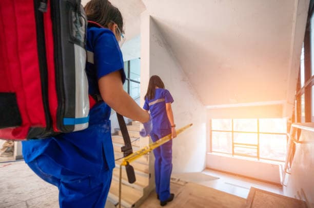 Two QuickCare emergency responders in blue uniforms ascending stairs in a workplace environment, carrying medical equipment with sunlight streaming through a window.