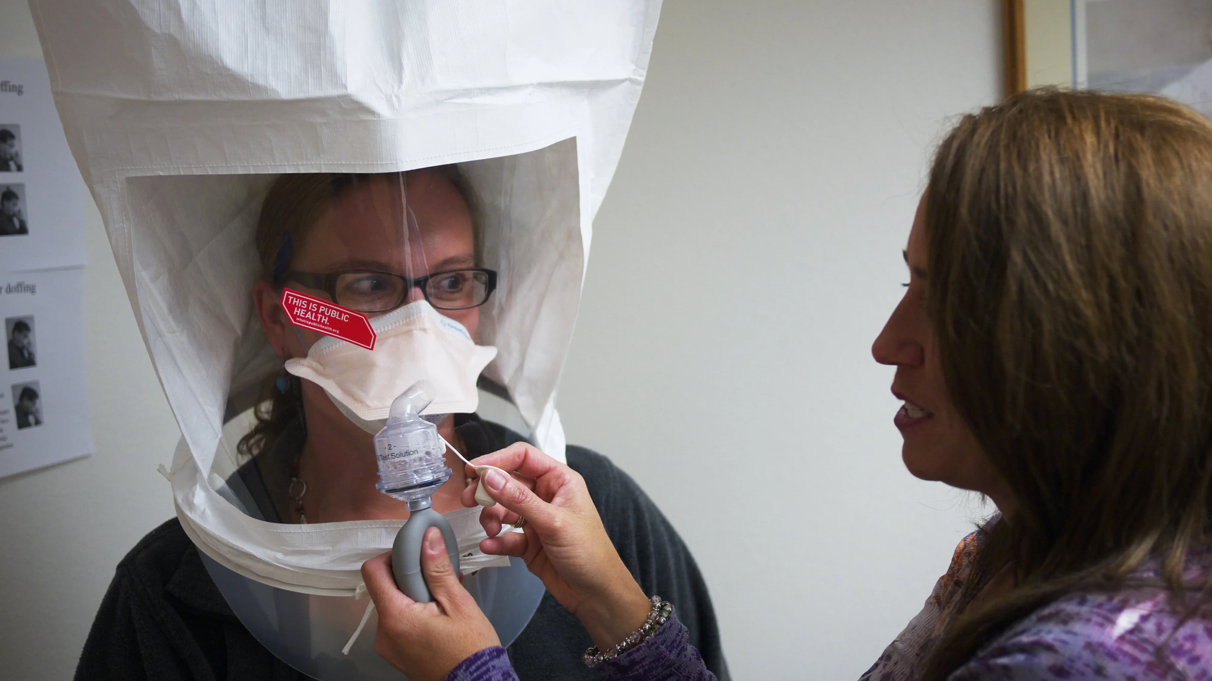 Respirator fit testing procedure for workplace safety, featuring a qualitative fit test with a healthcare professional ensuring proper mask seal under a hood.